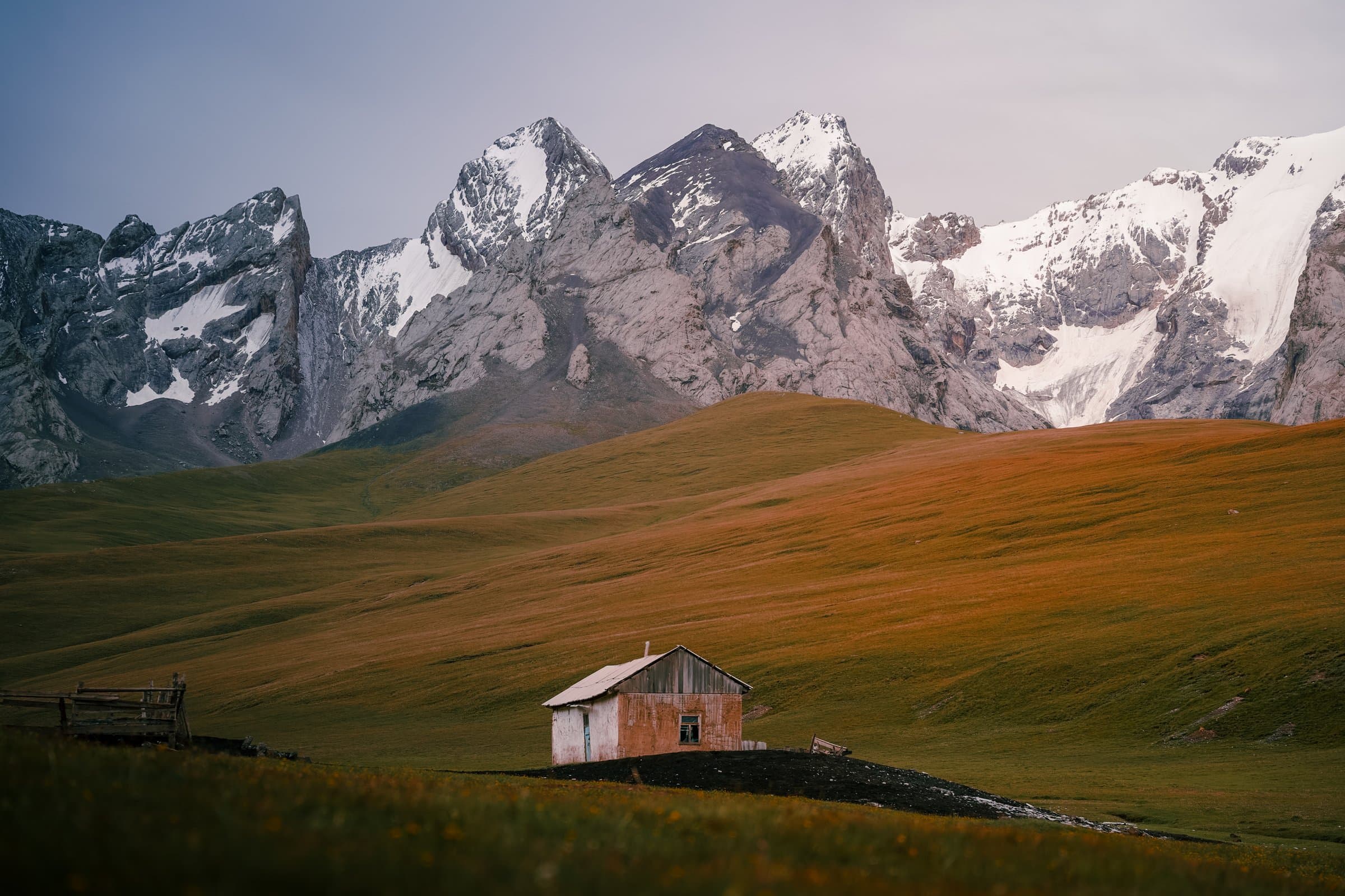 A wooden herder's hut in a golden valley below jagged, snow-streaked Kyrgyz peaks